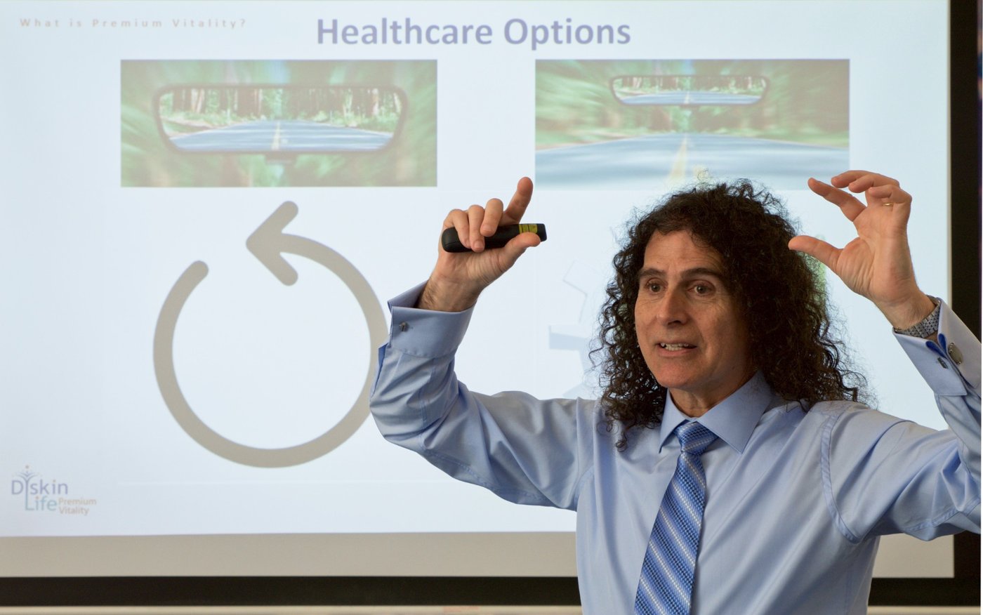 Dr Ari Diskin mid-presentation, hands raised as he explains a point, with his slides on the screen behind him