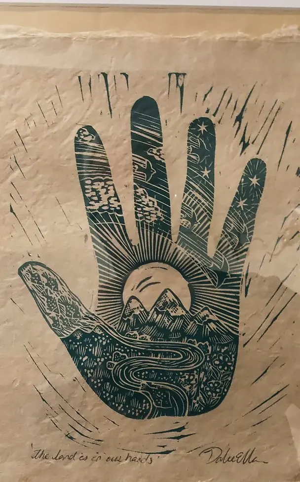 Linocut print of an open hand on textured paper, mountains and a rising sun set within the palm